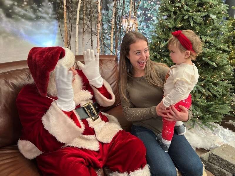 A woman holding a toddler sits next to a person dressed as Santa Claus on a brown couch, with a decorated Christmas tree and festive background behind them. The toddler looks at Santa, who is raising his hands.