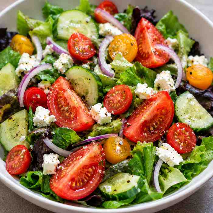 A fresh salad with lettuce, sliced cucumbers, cherry tomatoes, red onion, yellow tomatoes, and crumbled feta cheese, seasoned with black pepper, served in a white bowl.