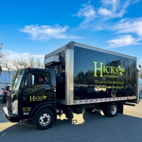 A black box truck with Hicks Nurseries and address printed on the side is parked outside on a sunny day. A person is visible in the driver’s seat. Blue sky and some trees are in the background.