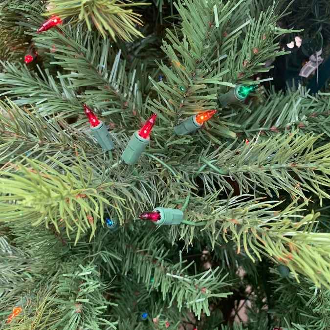 Small red, green, and orange string lights nestled in the needles of an artificial Christmas tree.